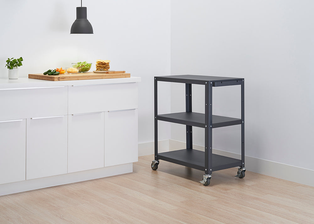 A kitchen scene shows white cabinets with minimalist handles and a dark grey, three-tiered rolling utility cart. On the counter, a wooden cutting board holds ingredients for tacos or similar food, illuminated by an overhead light.