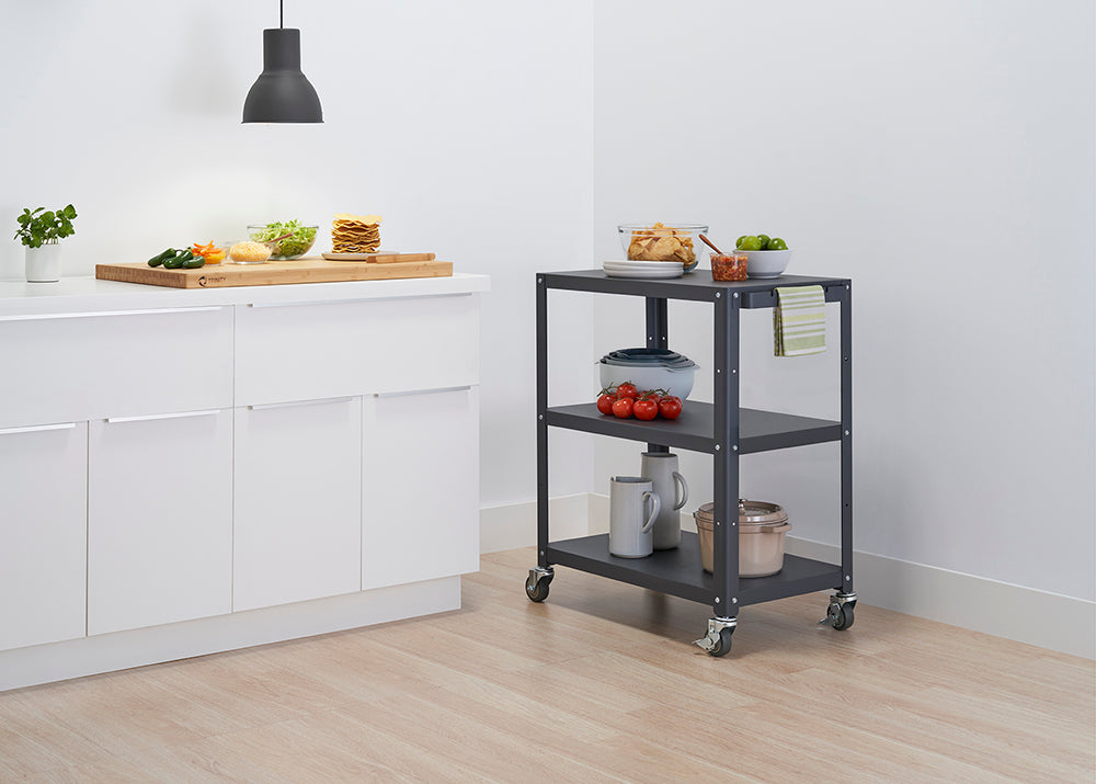 A dark grey, three-tiered metal utility cart on wheels stands next to white kitchen cabinets. The cart holds snacks like chips, limes, and tomatoes, as well as a stack of bowls and two mugs.