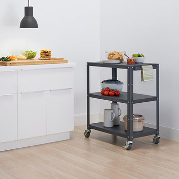 A dark grey, three-tiered metal utility cart on wheels stands next to white kitchen cabinets. The cart holds snacks like chips, limes, and tomatoes, as well as a stack of bowls and two mugs.