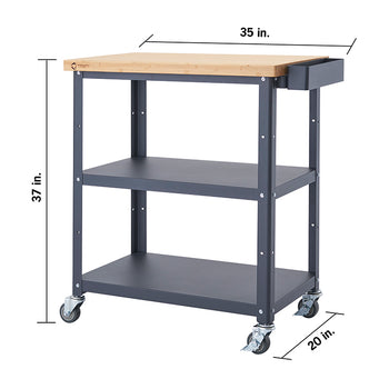 A front-facing view of a dark gray Trinity kitchen cart with a light brown bamboo top. The cart has two shelves below the top surface and is on four wheels, with dimensions of 37 inches high, 35 inches wide, and 20 inches deep.