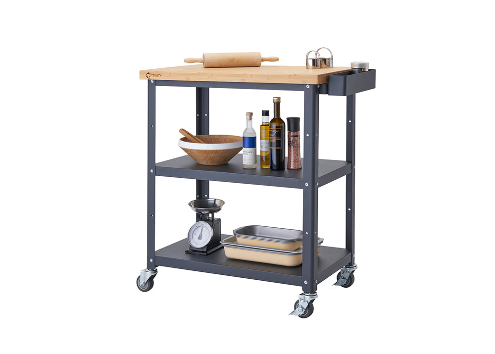 A dark gray Trinity 32" Bamboo Top Kitchen Cart on wheels is shown with a rolling pin on its bamboo top. The two lower shelves hold various kitchen items, including a bowl, bottles of oil, spices, a kitchen scale, and two baking pans.