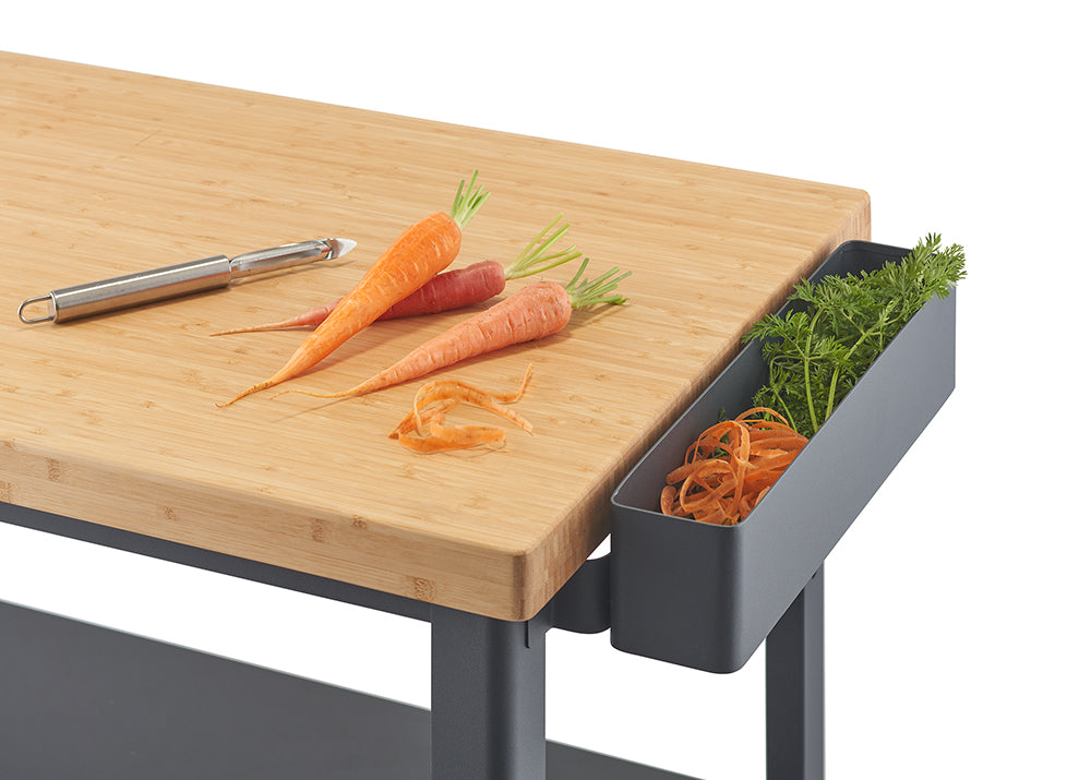 A close-up of a light wood countertop with three carrots, some carrot peelings, and a vegetable peeler on top. To the right, a gray bin attached to the counter holds carrot tops and more peelings.