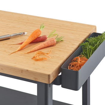 A close-up of a light wood countertop with three carrots, some carrot peelings, and a vegetable peeler on top. To the right, a gray bin attached to the counter holds carrot tops and more peelings.