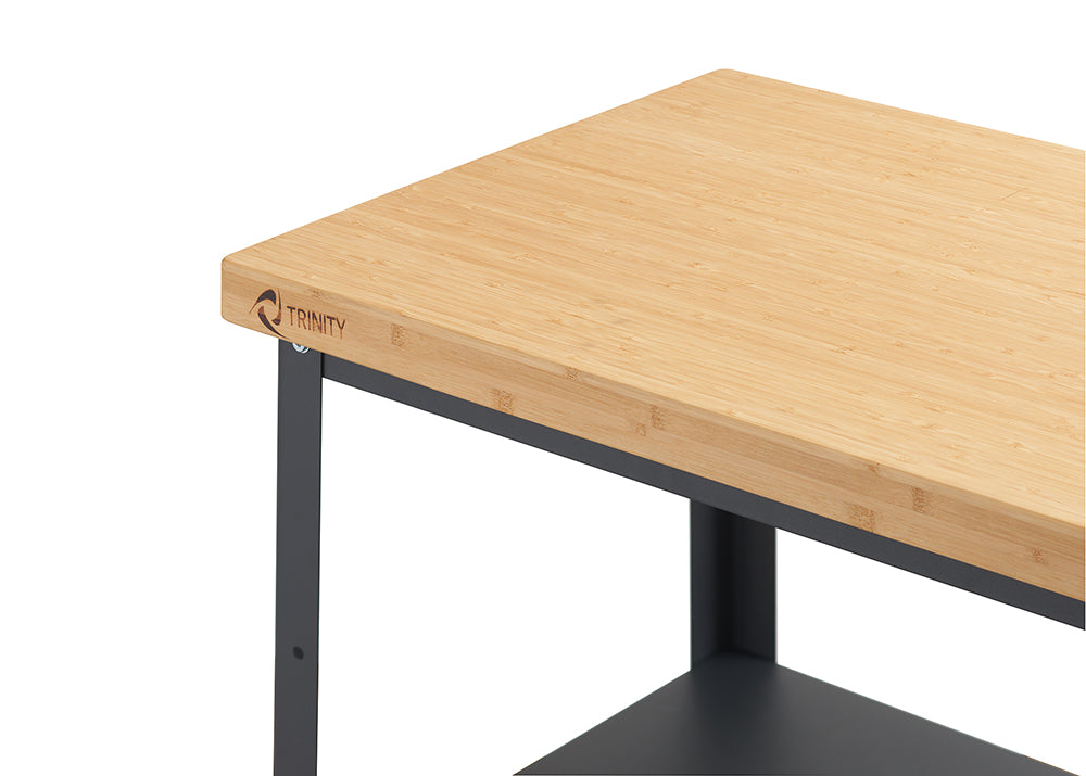 A close-up shot of the top right corner of a Trinity 32" Bamboo and Metal Kitchen Cart. The image highlights the natural bamboo countertop with the "TRINITY" logo subtly debossed or printed on the front left edge, and a dark metal frame supporting it.