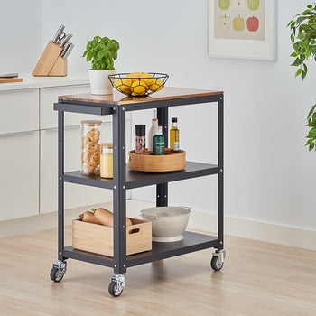 A dark gray metal and wood rolling kitchen cart is centrally placed in a modern kitchen. The cart has a wooden countertop holding a bowl of lemons and a potted herb, and two lower shelves organized with various kitchen items like jars of pasta, spices, oils, a wooden crate with squash, and a colander.