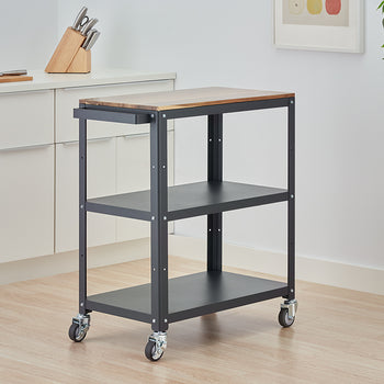 A three-tiered rolling kitchen cart with a dark gray metal frame and a wooden butcher block top stands on a light wood floor. The cart is positioned in a modern kitchen next to white cabinets, with a knife block and a cutting board visible on the counter.