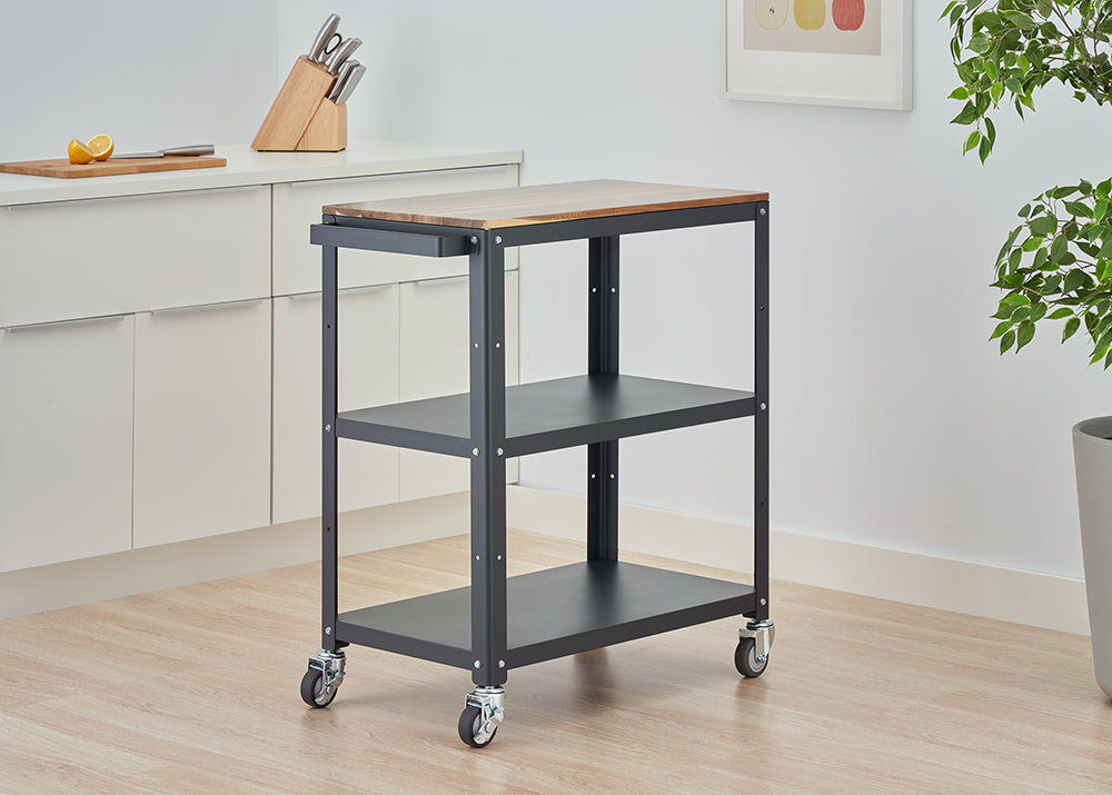 A three-tiered rolling kitchen cart with a dark gray metal frame and a wooden butcher block top stands on a light wood floor. The cart is positioned in a modern kitchen next to white cabinets, with a knife block and a cutting board visible on the counter.