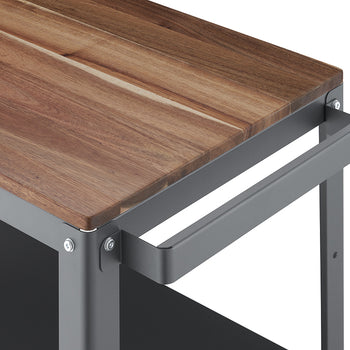 A close-up shot of a modern utility cart features a thick, wooden countertop with a natural wood grain texture. The countertop is supported by a gray metal frame, which includes a matching gray metal handle extending horizontally from the side.