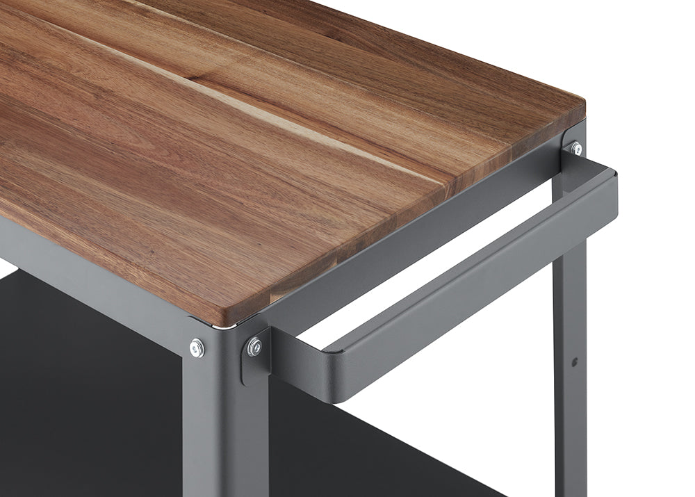 A close-up shot of a modern utility cart features a thick, wooden countertop with a natural wood grain texture. The countertop is supported by a gray metal frame, which includes a matching gray metal handle extending horizontally from the side.