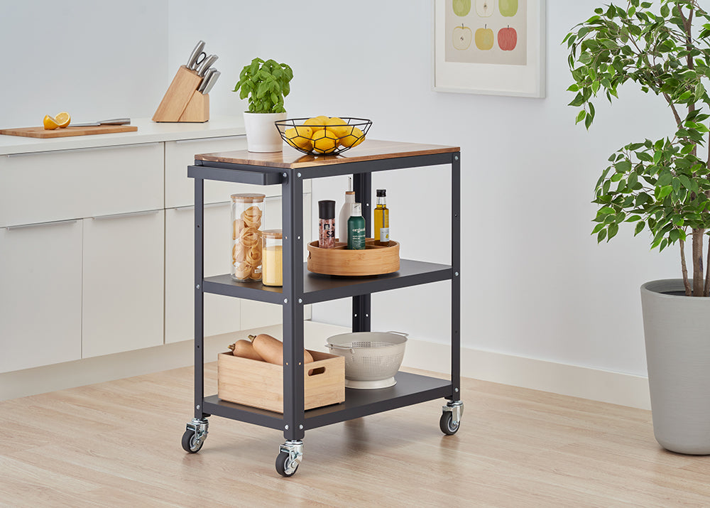 A dark gray metal and wood rolling kitchen cart is centrally placed in a modern kitchen. The cart has a wooden countertop holding a bowl of lemons and a potted herb, and two lower shelves organized with various kitchen items like jars of pasta, spices, oils, a wooden crate with squash, and a colander.