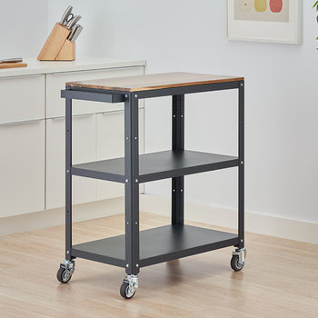 A three-tiered rolling kitchen cart with a dark gray metal frame and a wooden butcher block top stands on a light wood floor. The cart is positioned in a modern kitchen next to white cabinets, with a knife block and a cutting board visible on the counter.