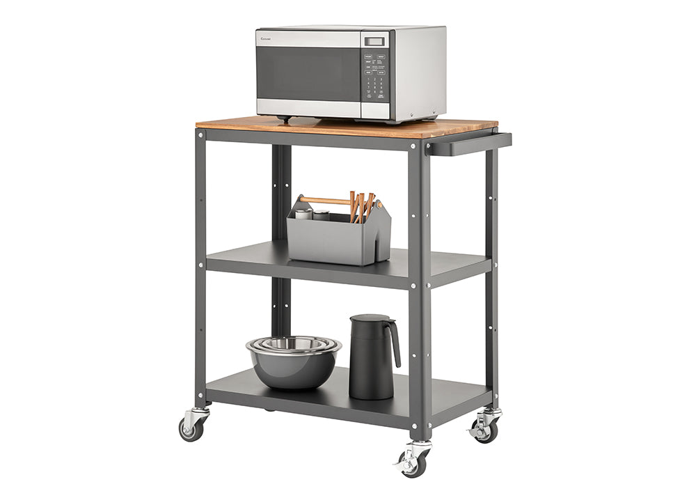 A three-tiered rolling utility cart with a dark gray metal frame and a light wood top shelf. A stainless steel microwave sits on the top, and the two lower shelves hold various kitchen items like bowls, a thermos, and a utensil caddy.