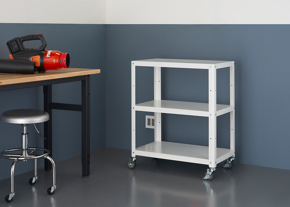 A white, three-tiered utility cart with wheels stands against a two-toned wall. To the left, a wooden table with a black metal frame holds a black and orange leaf blower. A chrome stool is tucked under the table.