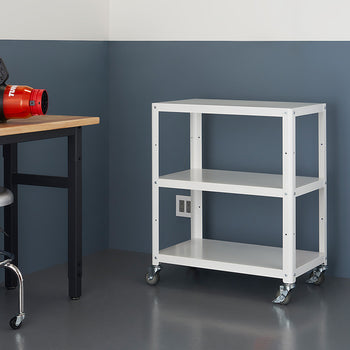 A white, three-tiered utility cart with wheels stands against a two-toned wall. To the left, a wooden table with a black metal frame holds a black and orange leaf blower. A chrome stool is tucked under the table.