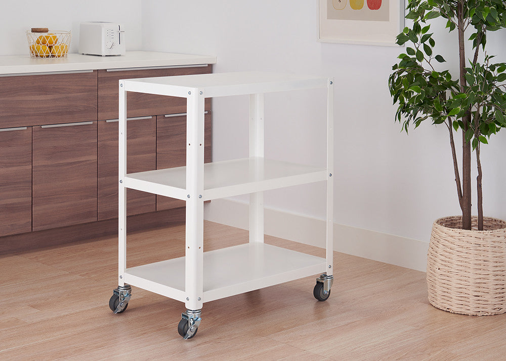 A white metal utility cart with three shelves on wheels is positioned in a kitchen setting against a light-colored wood floor. The cart is empty, with a white countertop and cabinets in the background, and a potted plant to the right.
