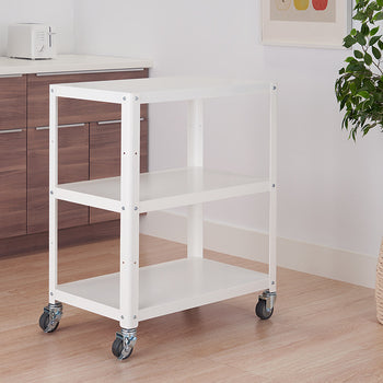 A white metal utility cart with three shelves on wheels is positioned in a kitchen setting against a light-colored wood floor. The cart is empty, with a white countertop and cabinets in the background, and a potted plant to the right.