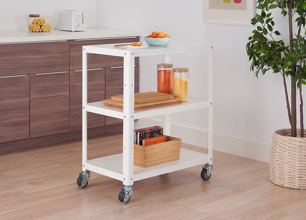 A white metal utility cart with three shelves on wheels sits in a kitchen setting. The cart holds various items, including jars, cutting boards, and a wooden box.