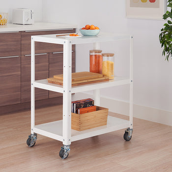 A white metal utility cart with three shelves on wheels sits in a kitchen setting. The cart holds various items, including jars, cutting boards, and a wooden box.