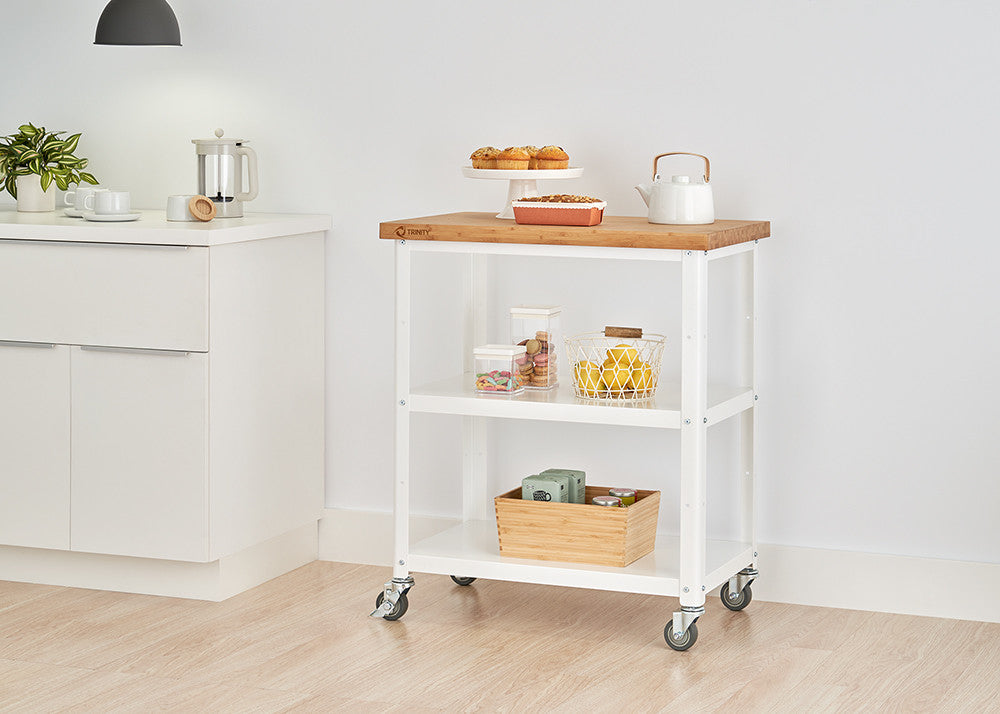 A white kitchen cart with a wooden top and two shelves is positioned in a kitchen setting against a white wall. The cart holds various kitchen items and is on wheels.