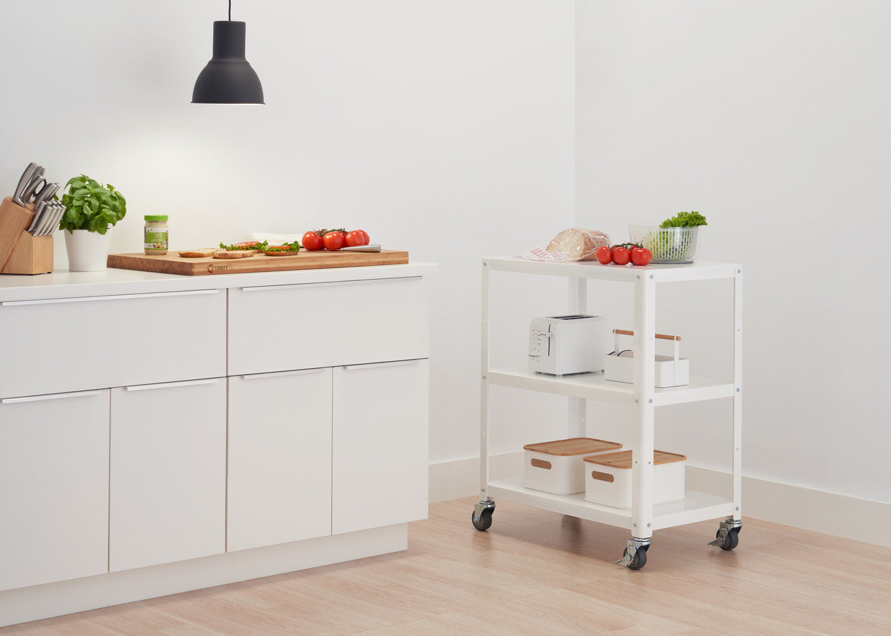 A bright, minimalist kitchen features white cabinets and a rolling white utility cart. A wooden cutting board with sliced tomatoes and bread sits on the countertop, illuminated by a hanging black pendant light.