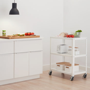 A bright, minimalist kitchen features white cabinets and a rolling white utility cart. A wooden cutting board with sliced tomatoes and bread sits on the countertop, illuminated by a hanging black pendant light.