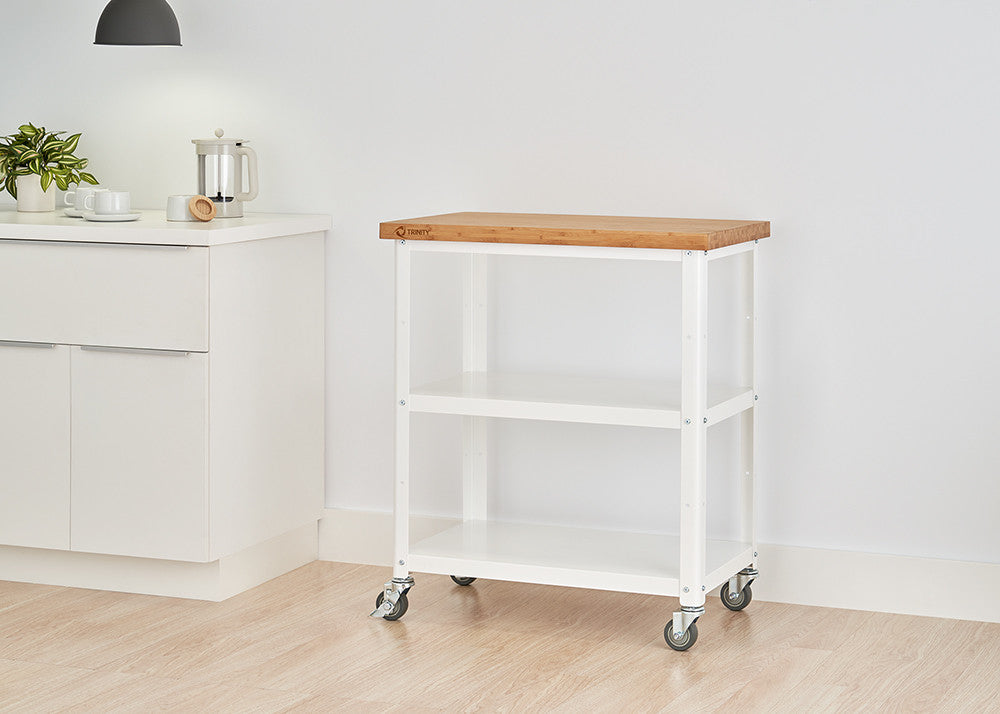 A white metal kitchen cart with a light wood butcher block top and two white wire shelves is shown against a white wall and light wood floor. The cart has four wheels and is positioned to the right of a white kitchen cabinet.