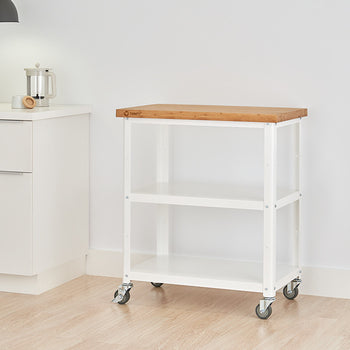 A white metal kitchen cart with a light wood butcher block top and two white wire shelves is shown against a white wall and light wood floor. The cart has four wheels and is positioned to the right of a white kitchen cabinet.