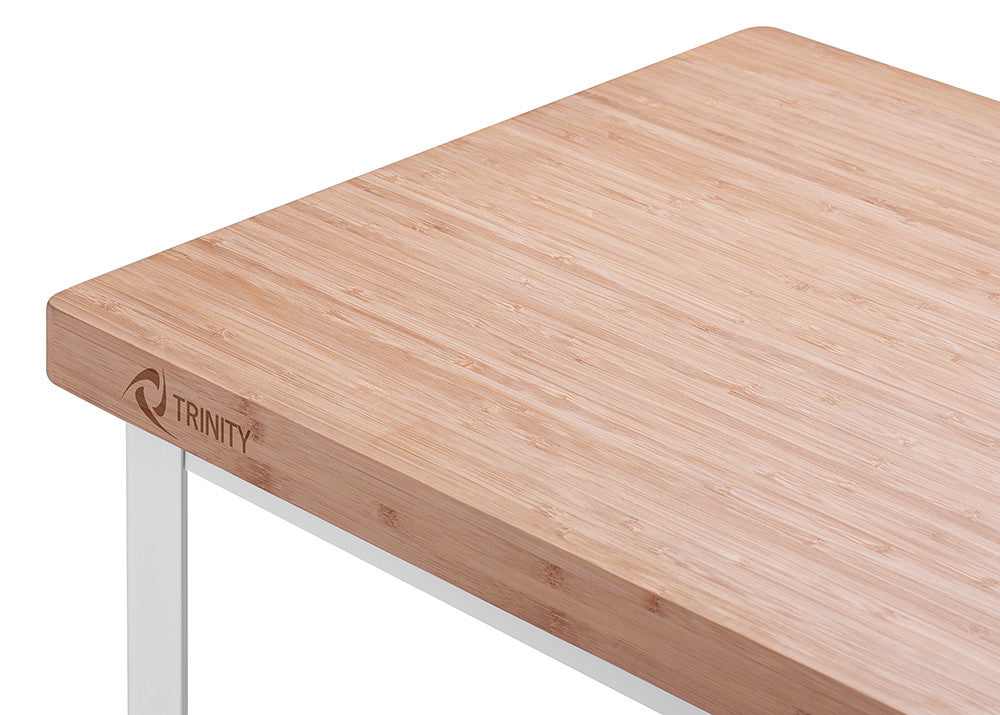 A close-up, angled shot shows the corner of a light bamboo butcher block countertop with the "TRINITY" logo engraved on the side. The countertop rests on a white metal frame, visible in the lower left corner.
