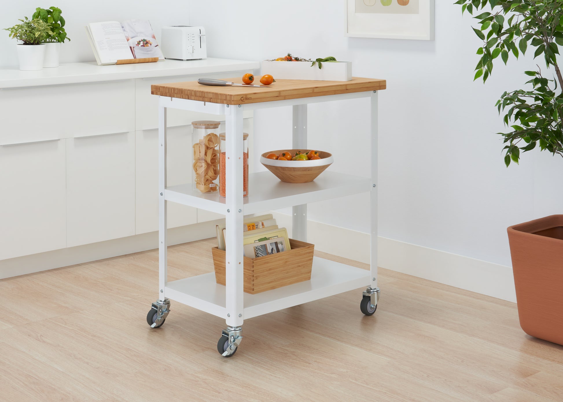 A Trinity 32" Bamboo Top Kitchen Cart with Side Rack is positioned in a bright kitchen. The cart has a wooden top, white shelves holding various items, and wheels.