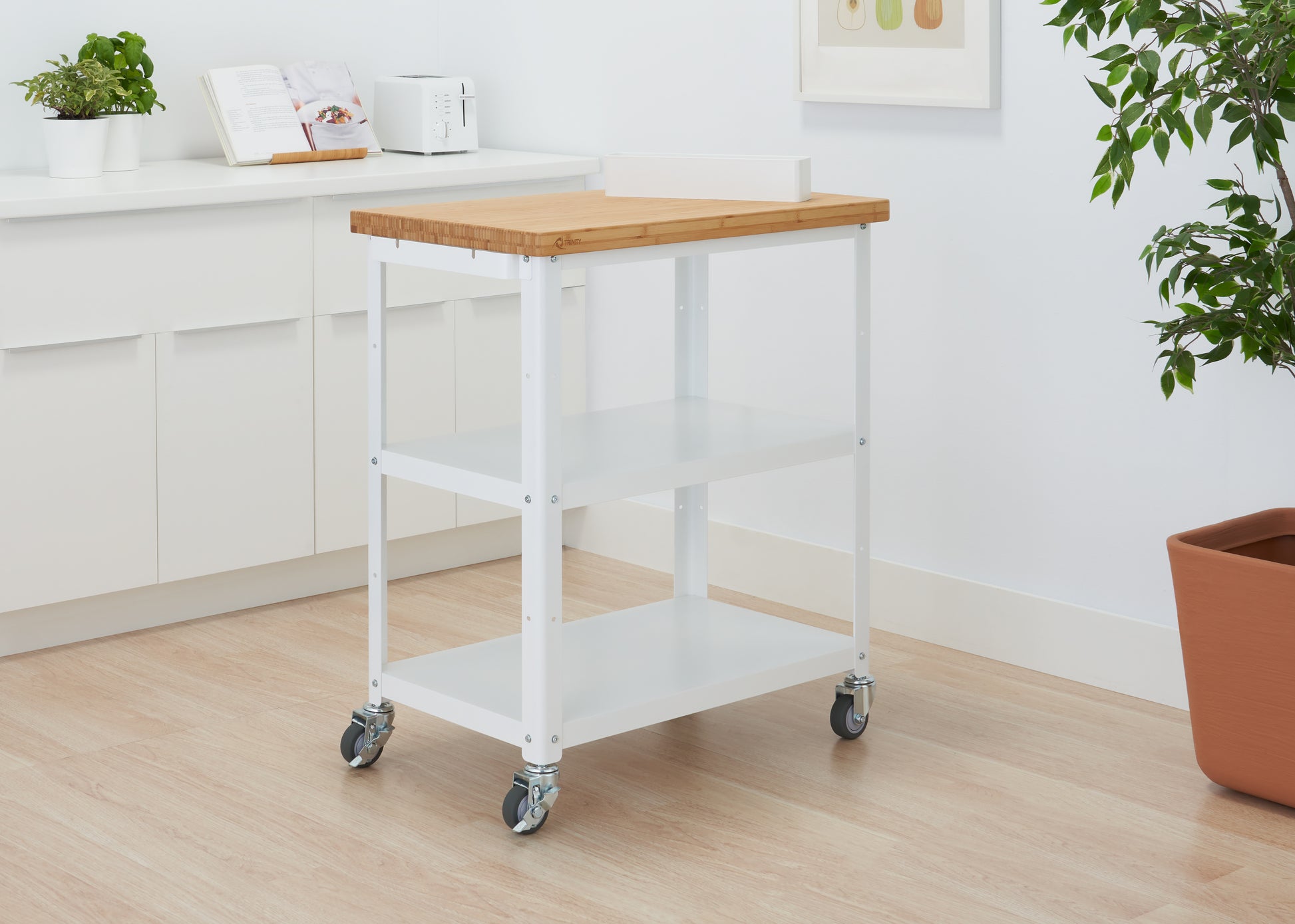 A white Trinity Basics 32" Bamboo Top Kitchen Cart with two shelves and wheels stands on a light wood floor in a brightly lit room. A toaster and a plant are visible on a white counter in the background.