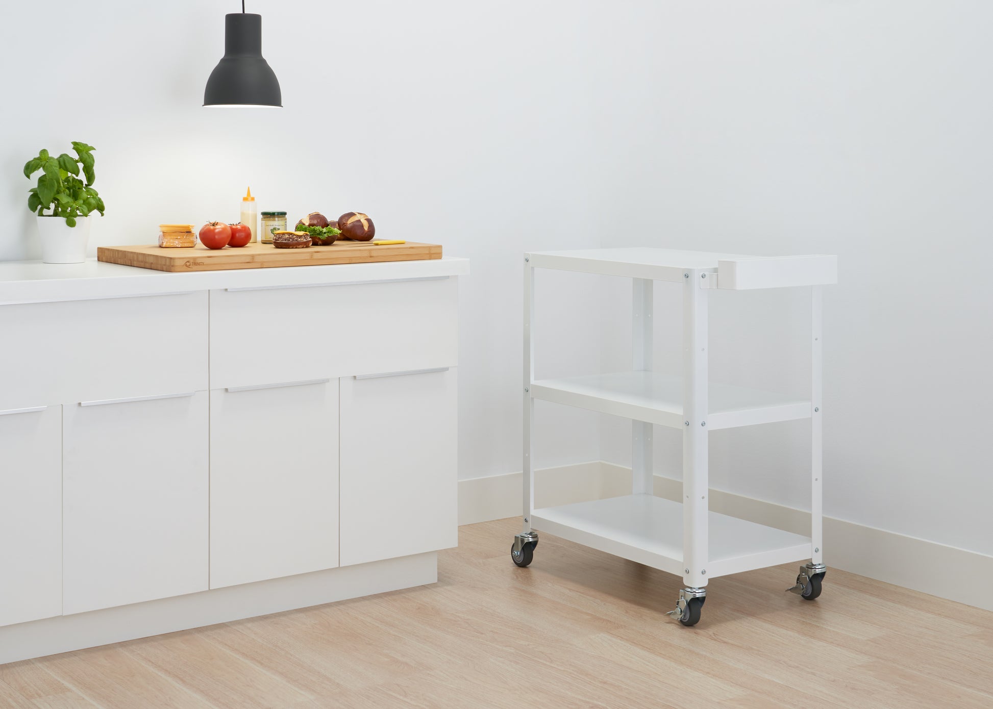 A white Trinity Metal Utility Cart with three shelves and wheels stands on a light wood floor. To the left, a white kitchen counter holds a wooden cutting board with tomatoes, onions, and other food items, illuminated by a black pendant light.