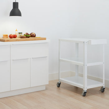 A white Trinity Metal Utility Cart with three shelves and wheels stands on a light wood floor. To the left, a white kitchen counter holds a wooden cutting board with tomatoes, onions, and other food items, illuminated by a black pendant light.