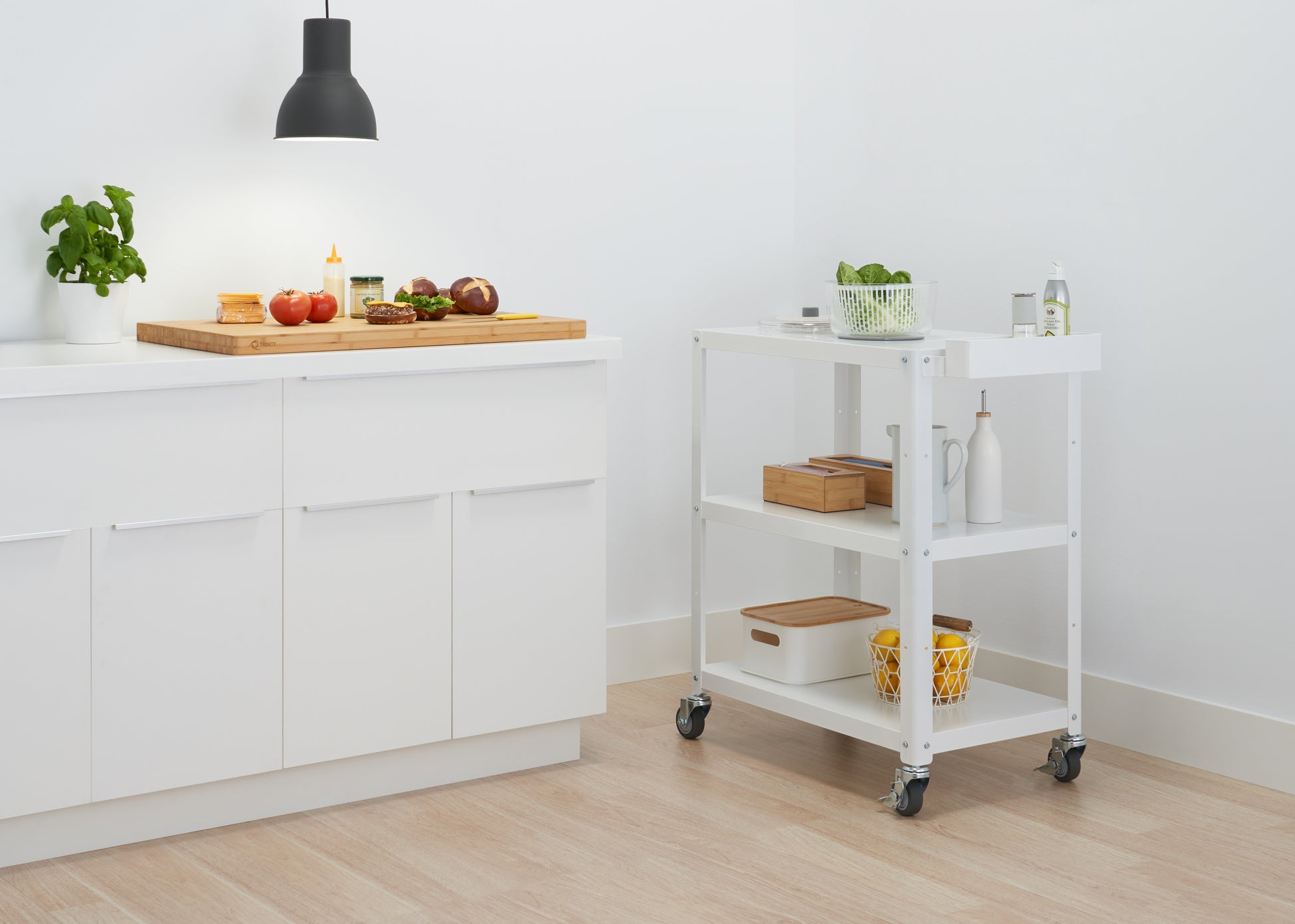 A minimalist kitchen features white cabinets with a light wood countertop, where a cutting board holds ingredients for a meal. To the right, a white utility cart on wheels offers additional storage for kitchen items.