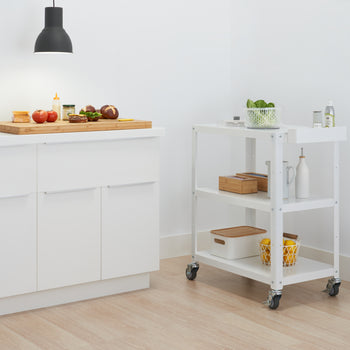 A minimalist kitchen features white cabinets with a light wood countertop, where a cutting board holds ingredients for a meal. To the right, a white utility cart on wheels offers additional storage for kitchen items.