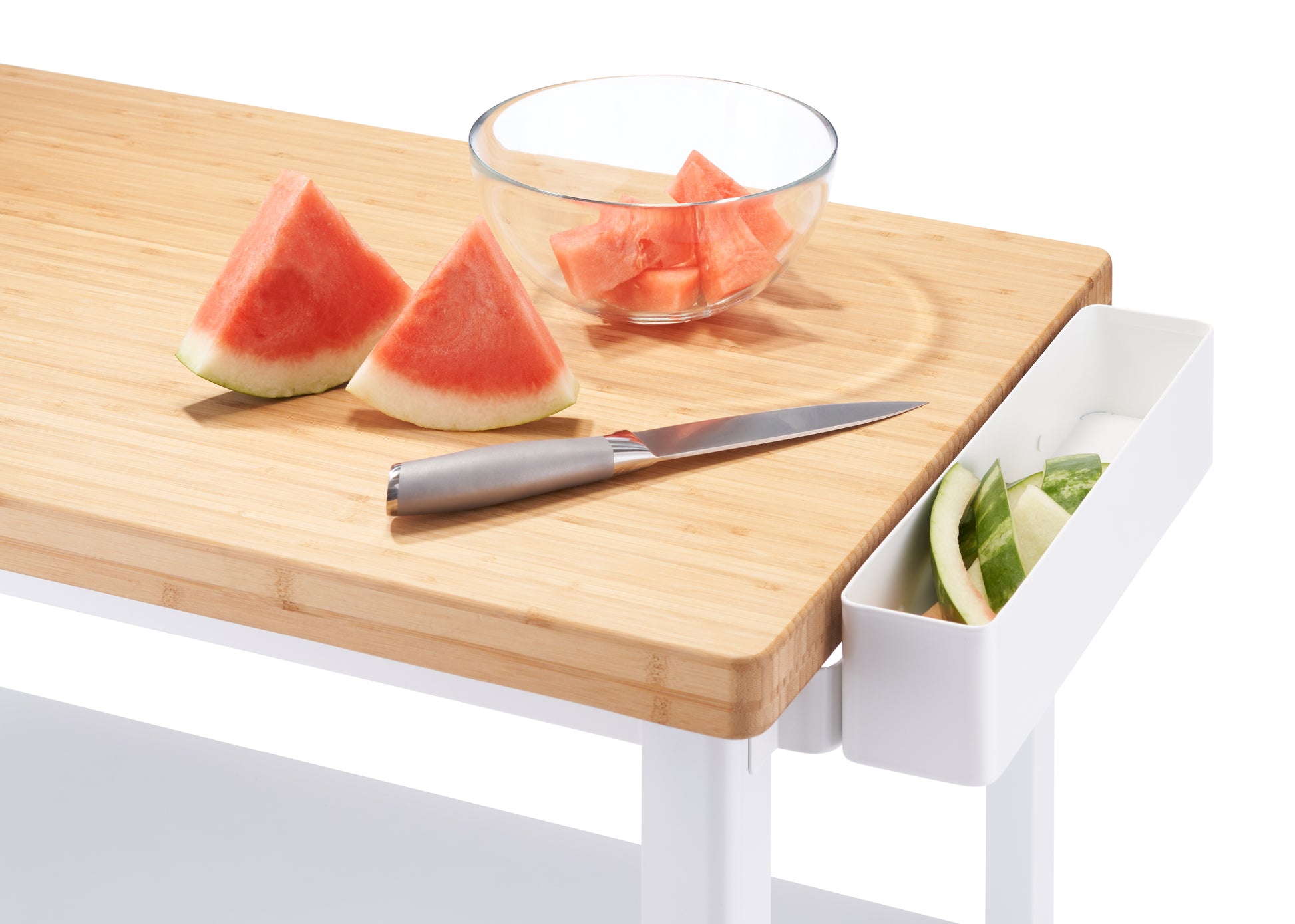 A white kitchen cart features a light wood top cutting board with two watermelon slices and a knife. To the right, a white container hangs from the board, holding watermelon rinds, while a glass bowl of cut watermelon sits behind the knife.