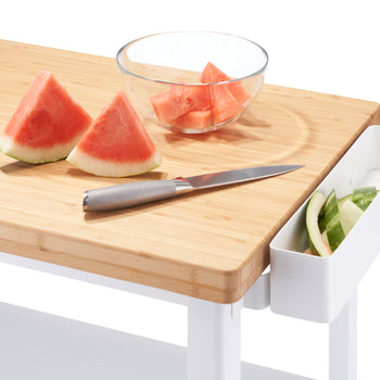 A white kitchen cart features a light wood top cutting board with two watermelon slices and a knife. To the right, a white container hangs from the board, holding watermelon rinds, while a glass bowl of cut watermelon sits behind the knife.