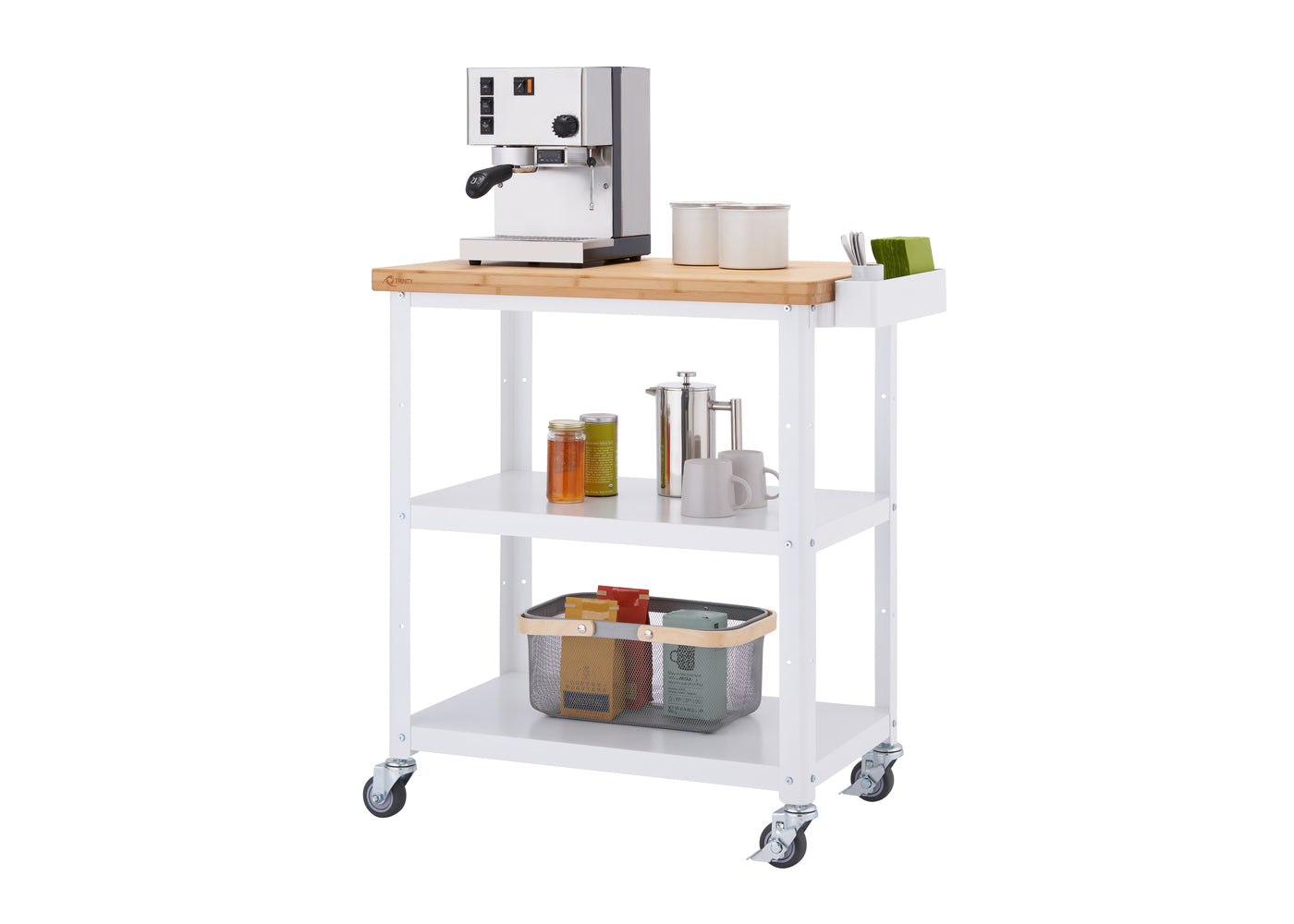 A white Trinity 32" Bamboo Top Kitchen Cart holds a variety of kitchen items. On the bamboo top, there's a stainless steel espresso machine, two cream-colored canisters, and a white organizer with green tea boxes. The two shelves below hold various items like jars, a French press, mugs, and a wire basket filled with colorful boxes.