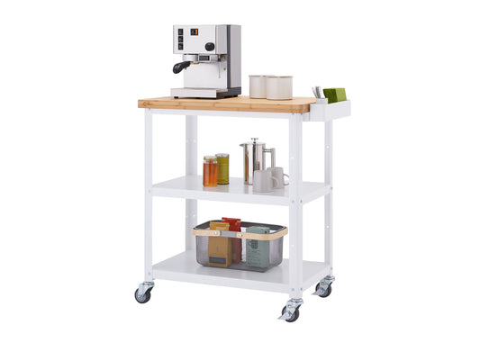 A white Trinity 32" Bamboo Top Kitchen Cart holds a variety of kitchen items. On the bamboo top, there's a stainless steel espresso machine, two cream-colored canisters, and a white organizer with green tea boxes. The two shelves below hold various items like jars, a French press, mugs, and a wire basket filled with colorful boxes.