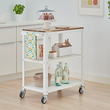 A white, three-tiered kitchen utility cart with a wooden top is shown on casters, positioned in a light-filled kitchen. The cart holds a cake under a dome, bottled water, a pot, and other kitchen accessories.