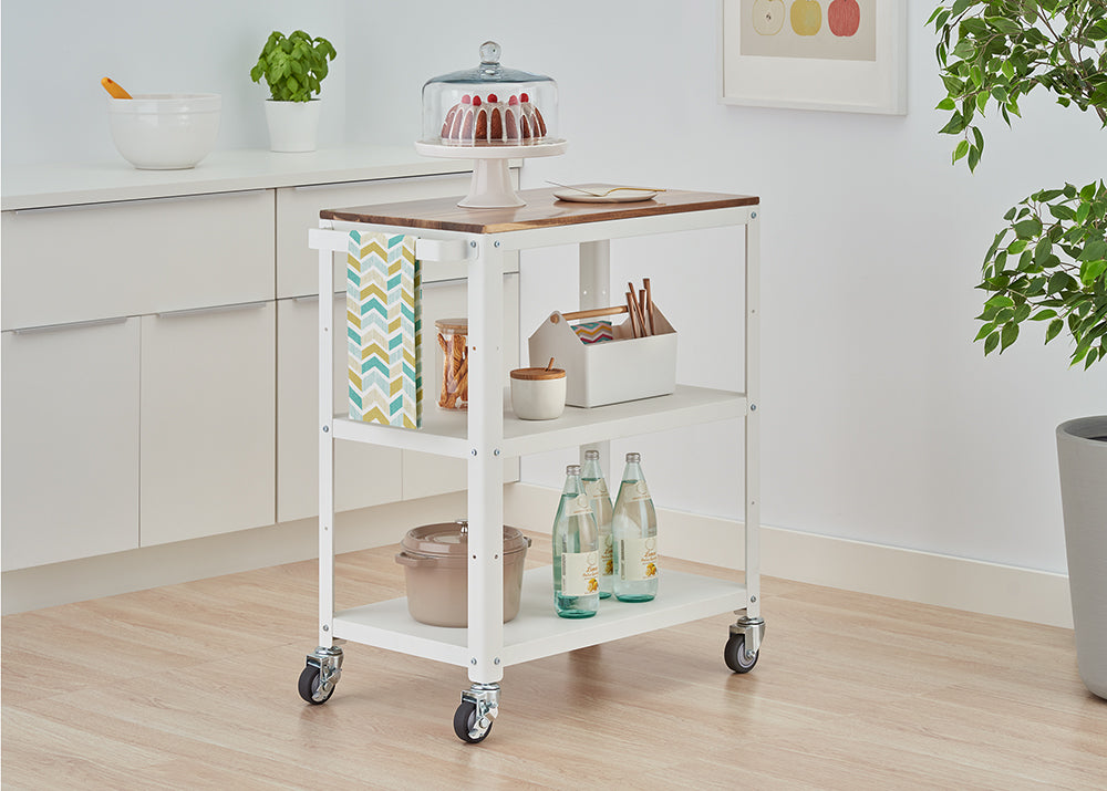 A white, three-tiered kitchen utility cart with a wooden top is shown on casters, positioned in a light-filled kitchen. The cart holds a cake under a dome, bottled water, a pot, and other kitchen accessories.