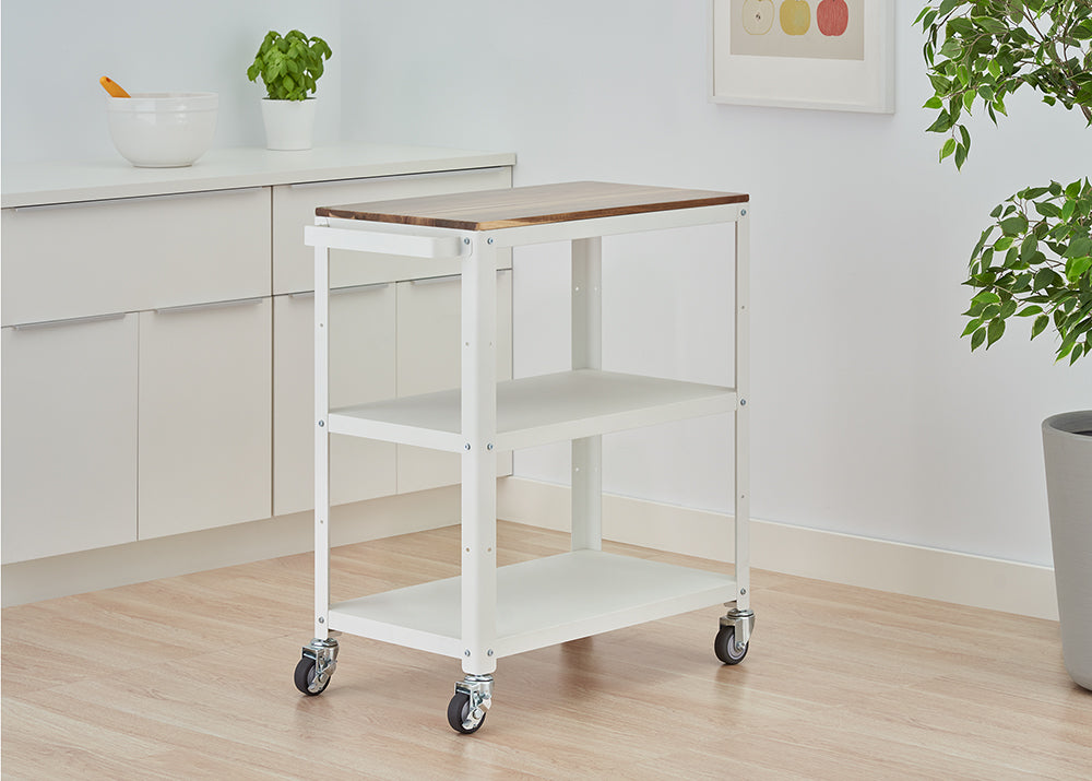 A white, three-tiered kitchen cart on wheels, featuring a wooden butcher block top. The cart is positioned on a light wooden floor in a bright room, next to white kitchen cabinets and some potted plants.