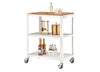 A white, three-tiered metal utility or bar cart with wheels is shown, featuring a wooden top surface. The cart is styled with bar accessories, including a gold cocktail tool set, a copper ice bucket, syrup bottles, and a four-pack of ginger beer.