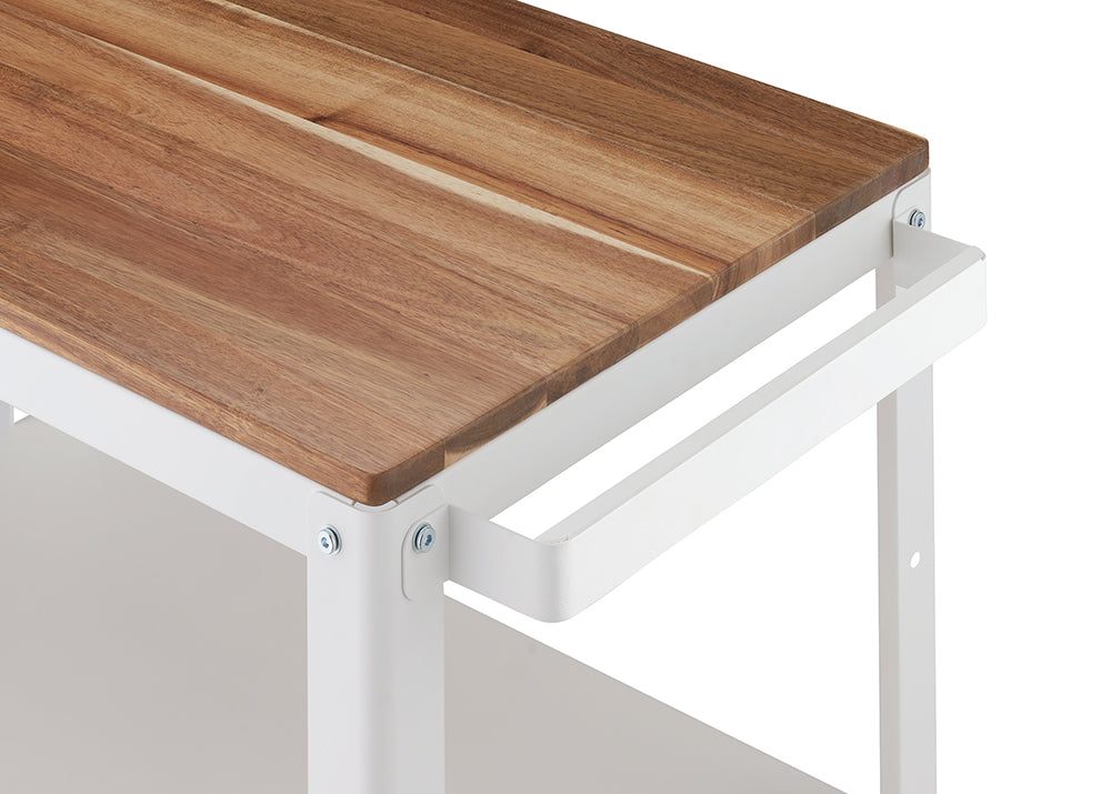 A close-up shot of the corner of a kitchen cart or utility table featuring a warm, butcher-block style wooden top set on a white metal frame. A simple white metal handle is attached to the side of the frame, near the edge of the wood top.