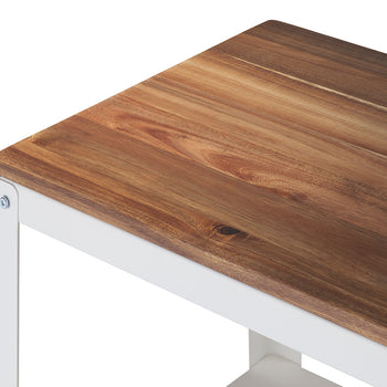 A close-up shot shows the corner of a table or bench featuring a warmly colored, richly grained wooden top, likely acacia or teak. The wood rests on a crisp white, painted metal frame, joined with visible screws.
