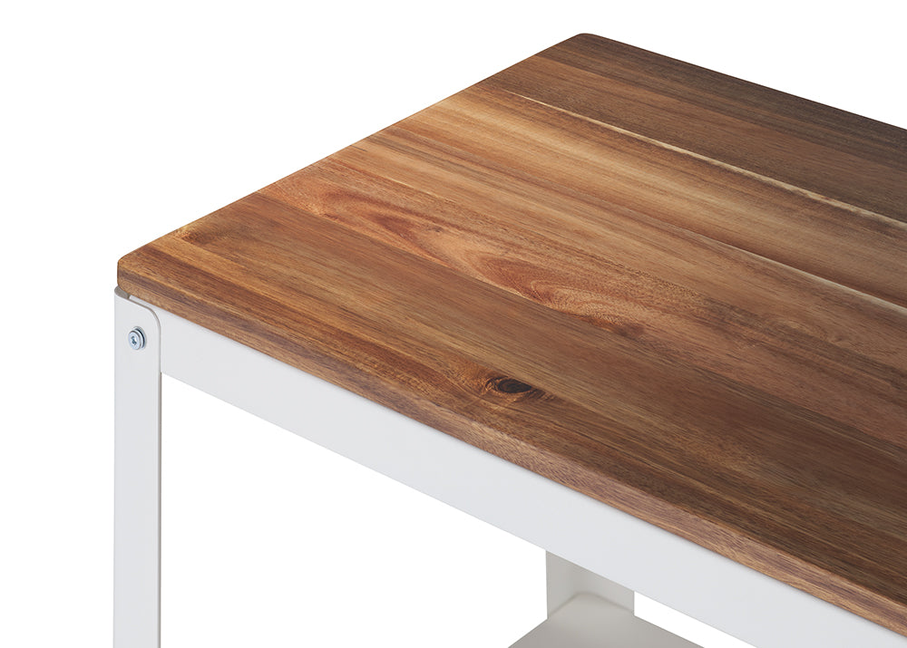 A close-up shot shows the corner of a table or bench featuring a warmly colored, richly grained wooden top, likely acacia or teak. The wood rests on a crisp white, painted metal frame, joined with visible screws.