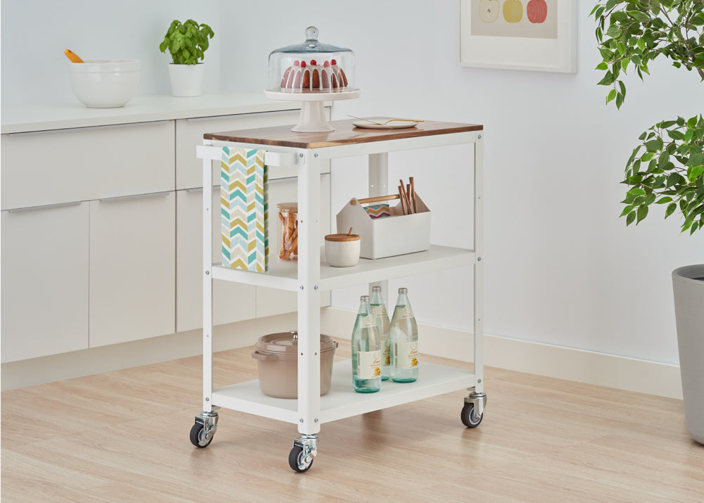 A white, three-tiered kitchen utility cart with a wooden top is shown on casters, positioned in a light-filled kitchen. The cart holds a cake under a dome, bottled water, a pot, and other kitchen accessories.