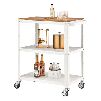 A white, three-tiered metal utility or bar cart with wheels is shown, featuring a wooden top surface. The cart is styled with bar accessories, including a gold cocktail tool set, a copper ice bucket, syrup bottles, and a four-pack of ginger beer.