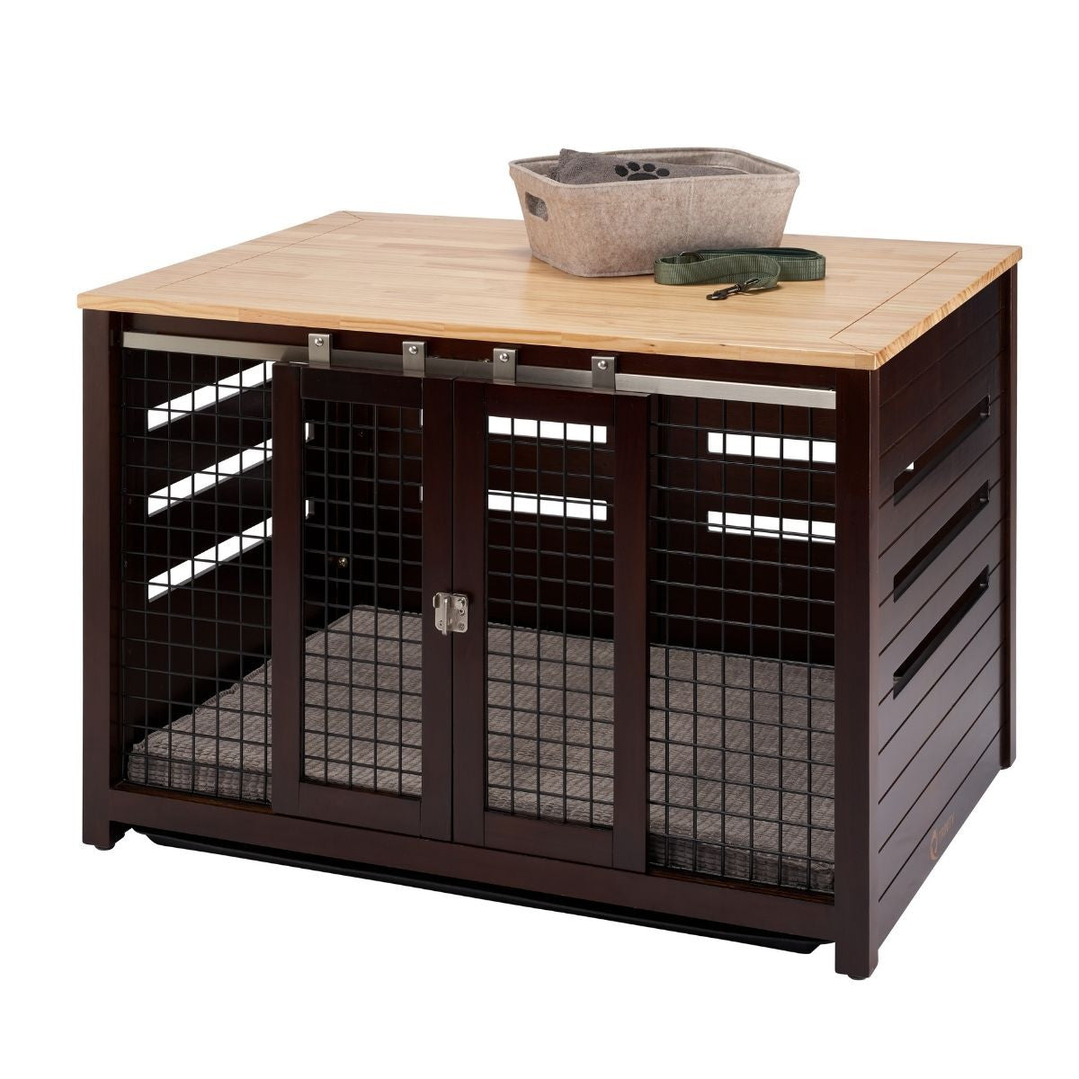 A square, dark brown pet crate with a light brown wooden top sits on a white background. The crate features sliding double doors with metal mesh and a soft gray mat inside, while a gray fabric basket and a green leash rest on its top surface.