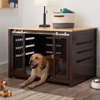 A golden Labrador retriever dog lies partially inside a dark brown pet crate that doubles as an accent table with a light wood top. The crate features sliding metal doors and is positioned on a light-colored wooden floor next to a woven basket and a floor lamp.