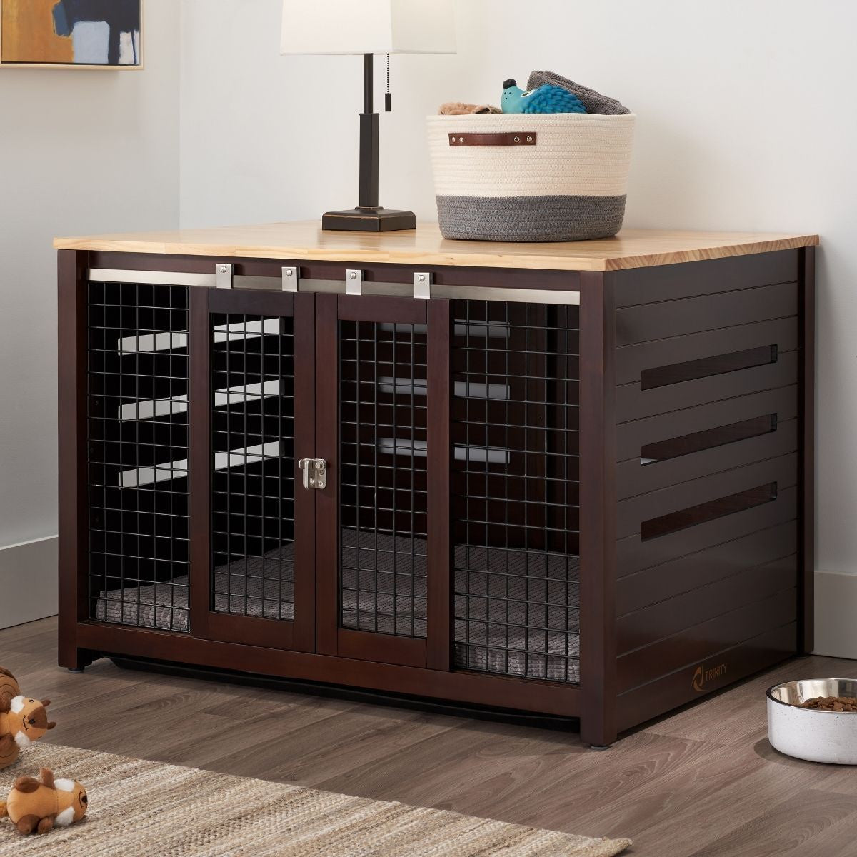 A dark brown wooden pet crate with a light brown wooden top sits on a light brown wooden floor against a white wall. The crate has two sliding metal mesh doors with a latch in the center, and a white lamp and a woven basket with blue and white items sit on top.